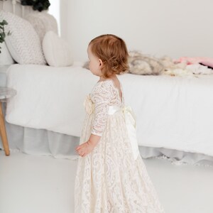 May include: A young girl wearing a white lace dress with a satin sash and a floral embellishment. The dress has long sleeves and a flowing skirt. The girl is standing in a white room with a bed in the background.