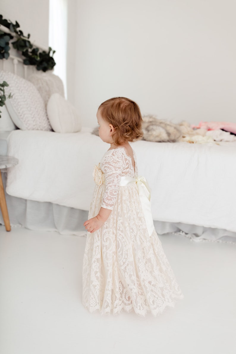May include: A young girl wearing a white lace dress with a satin sash and a floral embellishment. The dress has long sleeves and a flowing skirt. The girl is standing in a white room with a bed in the background.