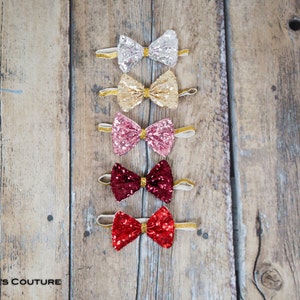 May include: Five sequined bow ties in different colors, including silver, gold, pink, burgundy, and red. The bow ties are arranged vertically on a wooden surface.