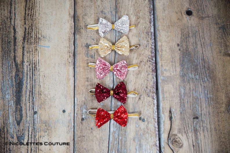 May include: Five sequined bow ties in different colors, including silver, gold, pink, burgundy, and red. The bow ties are arranged vertically on a wooden surface.