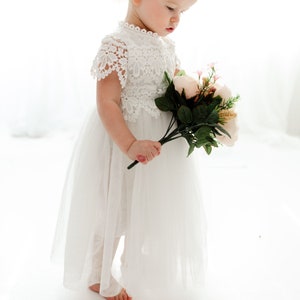 May include: A young girl in a white lace dress and a white headband with a bow holds a bouquet of pink and white flowers.