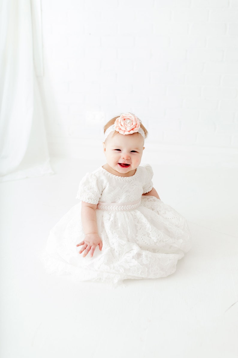 Puede incluir: Una ni&ntilde;a peque&ntilde;a con un vestido blanco de encaje y una diadema de flores rosas. Est&aacute; sentada sobre un fondo blanco y sonriendo.