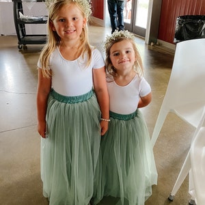 May include: Two young girls wearing white shirts and sage green tulle skirts. They are both wearing flower crowns and smiling at the camera.