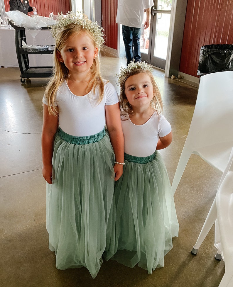 May include: Two young girls wearing white shirts and sage green tulle skirts. They are both wearing flower crowns and smiling at the camera.