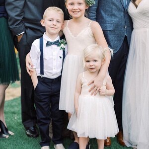 May include: Two young children, a boy and a girl, wearing formal attire for a wedding. The boy is wearing a white shirt, a navy blue bow tie, and navy blue suspenders. The girl is wearing a white tulle dress with lace trim. They are standing in front of a group of adults.