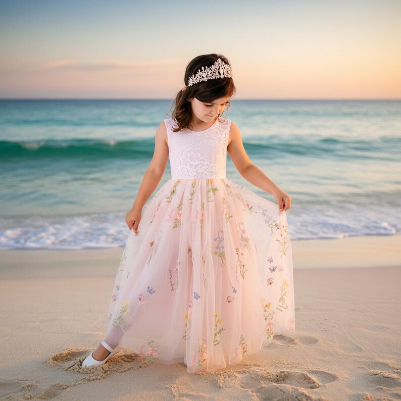 May include: A floor-length pale pink dress featuring a sleeveless lace bodice and a tulle skirt adorned with embroidered colourful floral patterns. The garment is paired with a silver-toned tiara with clear crystals worn on the head.