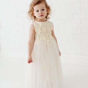 May include: A young girl wearing a white tulle dress with a cream lace bodice. The dress has a sleeveless design and a full skirt.