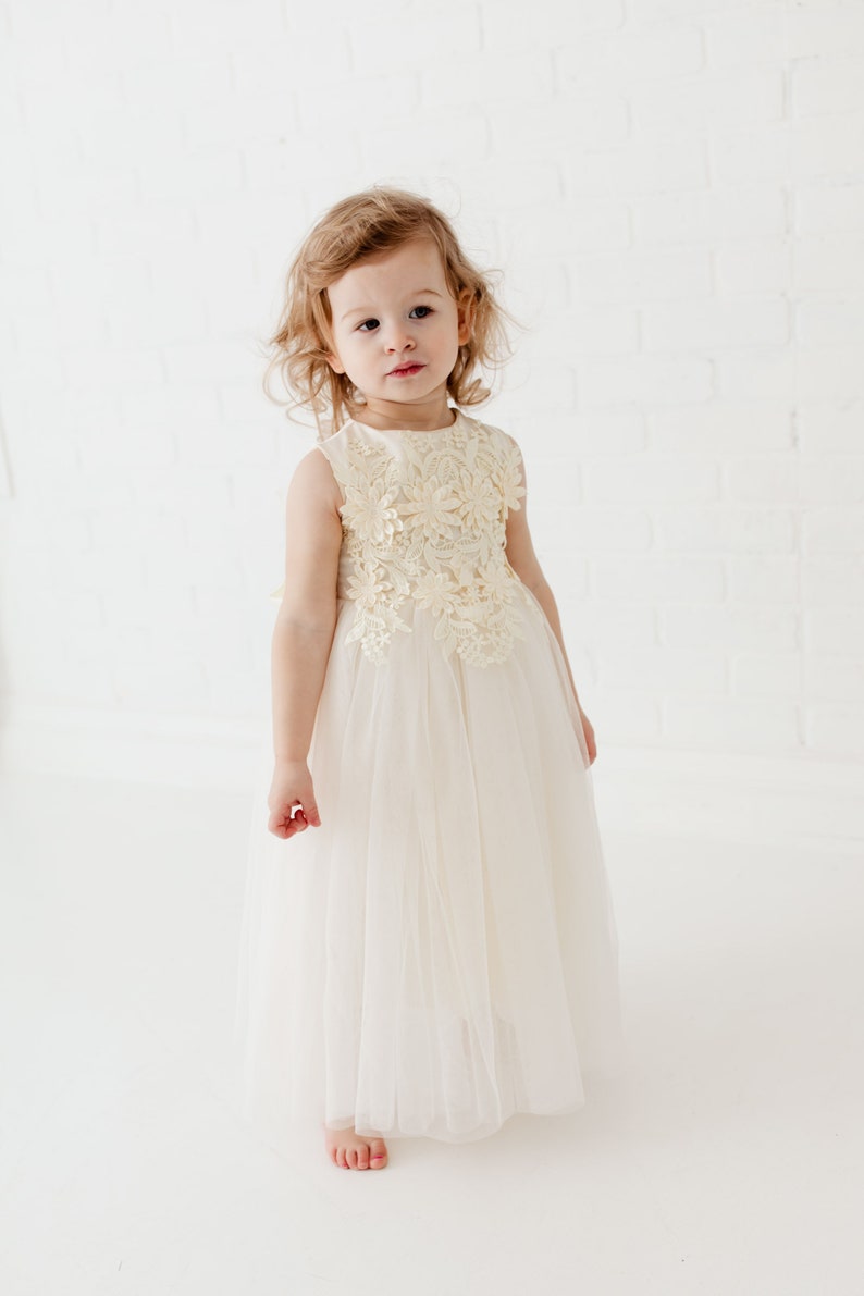 May include: A young girl wearing a white tulle dress with a cream lace bodice. The dress has a sleeveless design and a full skirt.