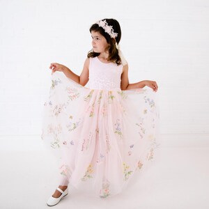 May include: A pale pink sleeveless dress featuring a bodice with white floral embroidery and a full tulle skirt adorned with colourful embroidered flowers and butterflies. The outfit includes a matching floral headband with white petals.