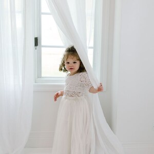 May include: A young girl in a white lace top and tulle skirt stands near a window with sheer white curtains. The outfit is a light, airy style, perfect for a special occasion. The room is bright and minimalist, with white walls and flooring.