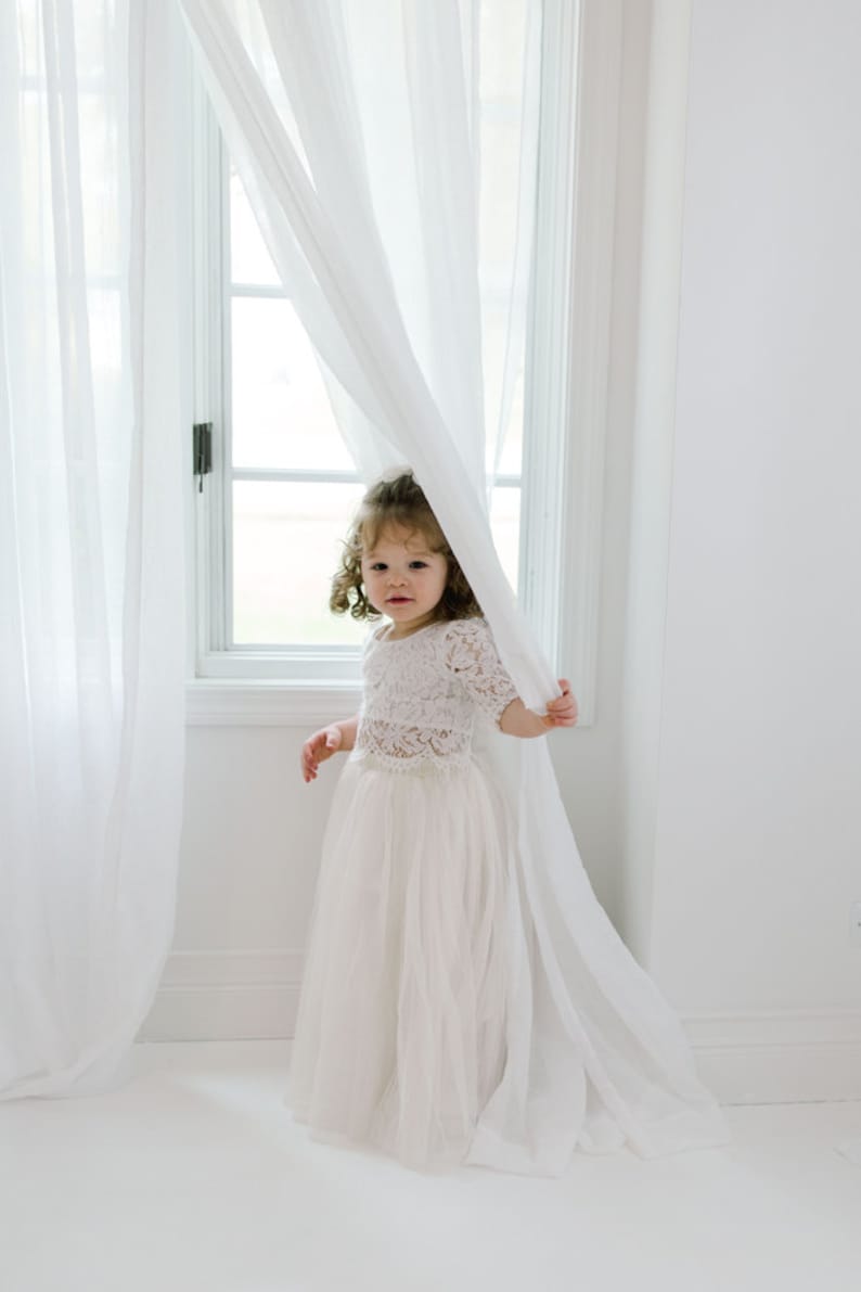 May include: A young girl in a white lace top and tulle skirt stands near a window with sheer white curtains. The outfit is a light, airy style, perfect for a special occasion. The room is bright and minimalist, with white walls and flooring.