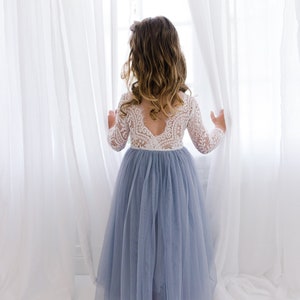 May include: A young girl in a light blue tulle dress with a white lace bodice stands in front of a window with white curtains. She is looking out the window.