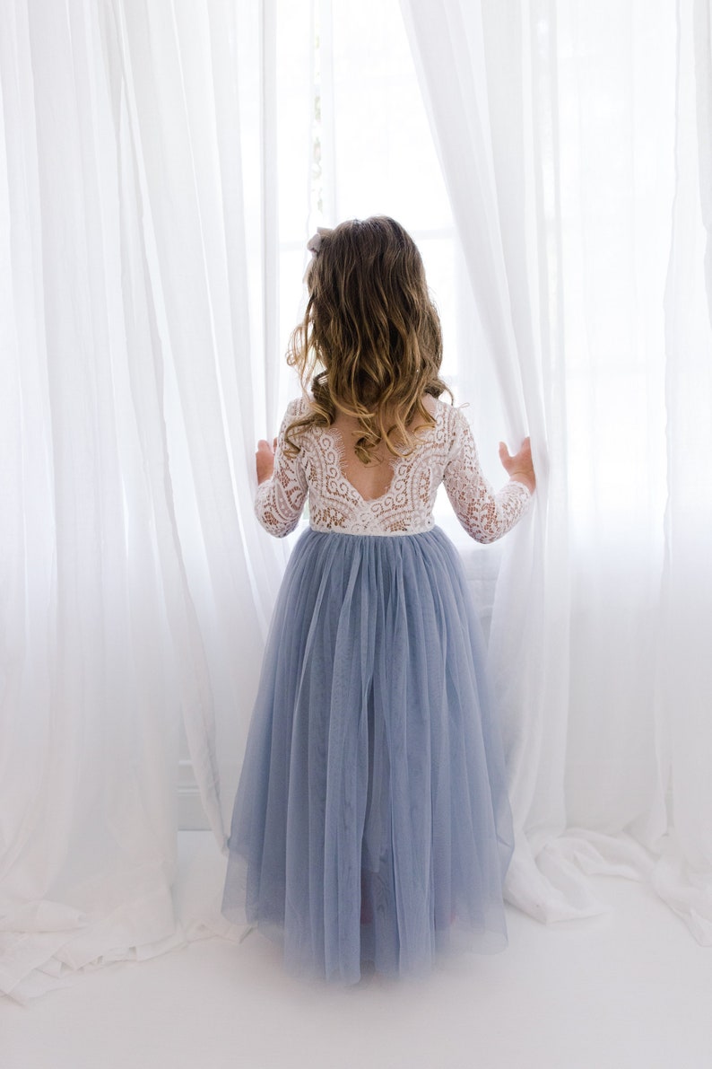 May include: A young girl in a light blue tulle dress with a white lace bodice stands in front of a window with white curtains. She is looking out the window.