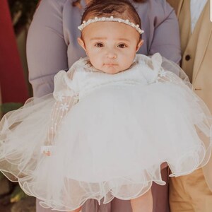 K&ouml;nnte beinhalten: Ein wei&szlig;es Babykleid mit einem T&uuml;llrock und Spitzen&auml;rmeln. Das Kleid hat ein Perlenhaarband.