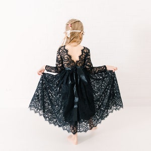 May include: A long-sleeved black lace dress featuring a deep V-shaped back, a scalloped hemline, and a wide satin ribbon tied into a bow at the waist. The intricate floral lace pattern covers the bodice and the full, flowing skirt of the garment.