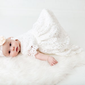 K&ouml;nnte beinhalten: Ein Baby in einem wei&szlig;en Spitzenkleid und einem Stirnband mit einer Blume liegt auf einem wei&szlig;en, flauschigen Teppich.