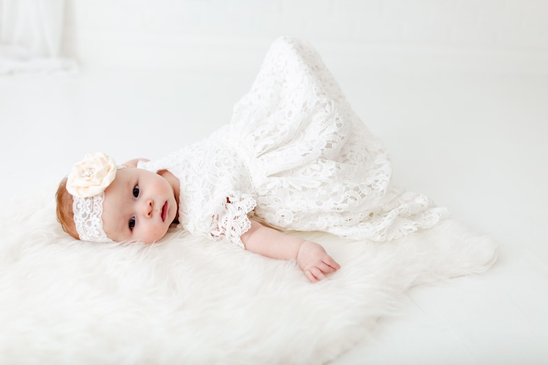 K&ouml;nnte beinhalten: Ein Baby in einem wei&szlig;en Spitzenkleid und einem Stirnband mit einer Blume liegt auf einem wei&szlig;en, flauschigen Teppich.
