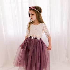 May include: A young girl wearing a white lace top and a long, dusty rose tulle skirt. She is wearing a burgundy bow headband.