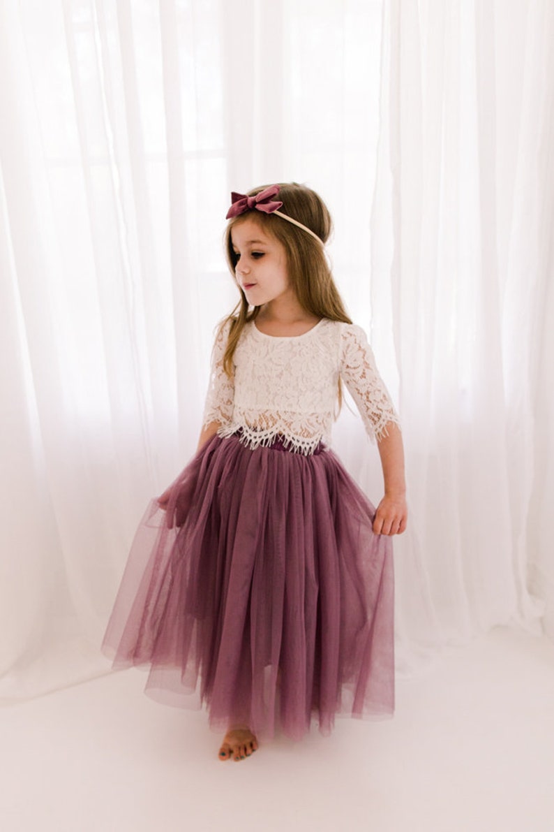 May include: A young girl wearing a white lace top and a long, dusty rose tulle skirt. She is wearing a burgundy bow headband.
