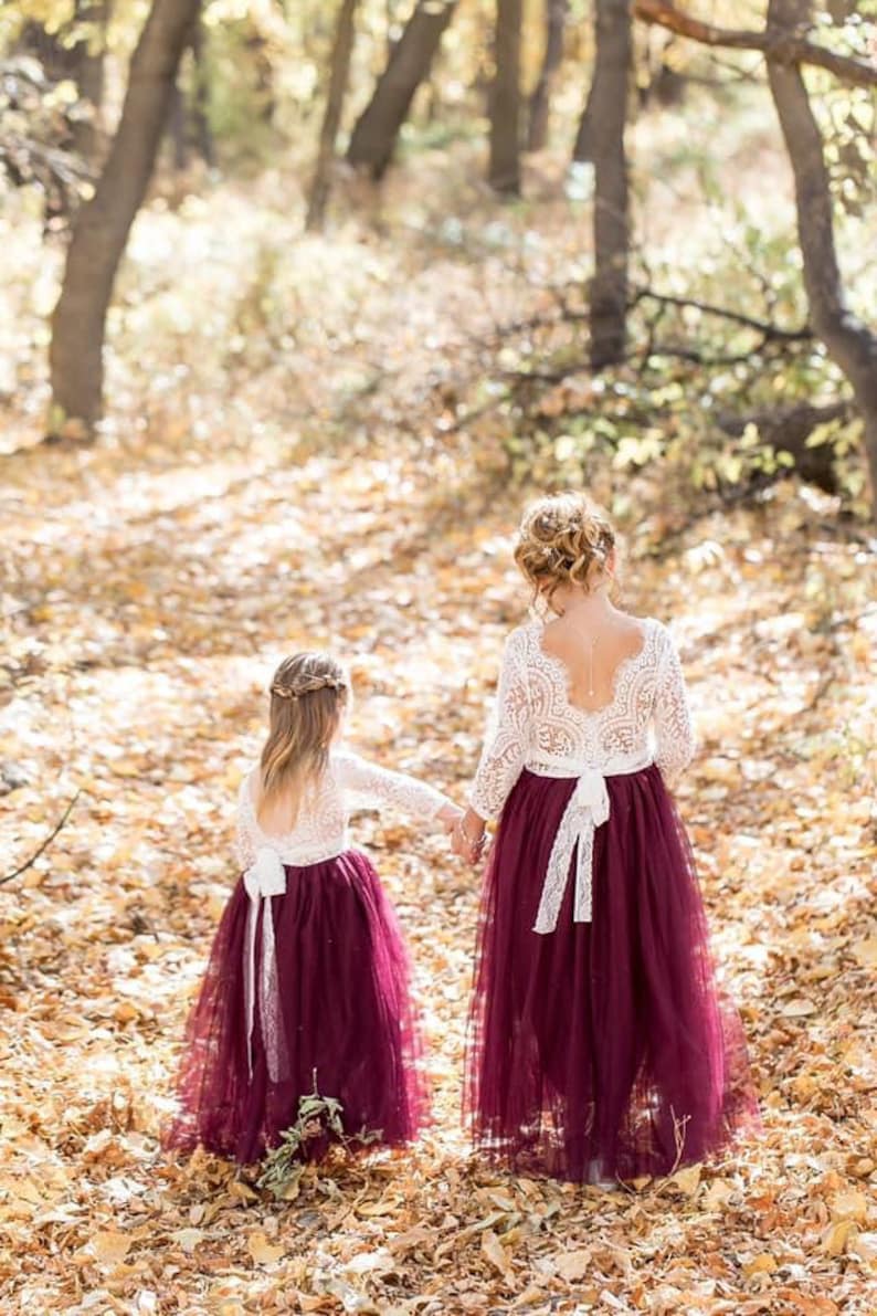 May include: Two dresses featuring long-sleeved white lace bodices with open back designs and floor-length, deep burgundy tulle skirts. Each dress includes a white lace tie at the waist, creating a cohesive look with matching textures and colors.