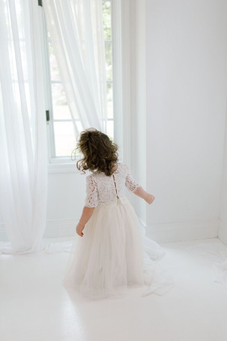 May include: A young child wearing a white lace dress with a tulle skirt, standing near a window with sheer white curtains. The dress has short sleeves and a detailed lace bodice.