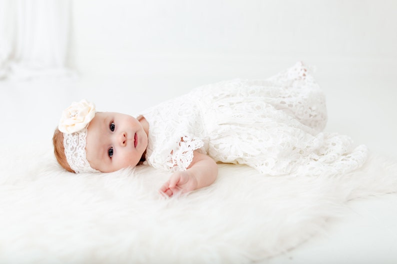 May include: A baby wearing a white lace dress and a headband with a flower on it. The baby is lying on a white fluffy surface.