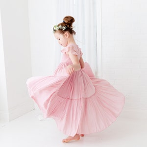 May include: A young girl wearing a pink, tiered dress with a floral crown spins around in a white room.