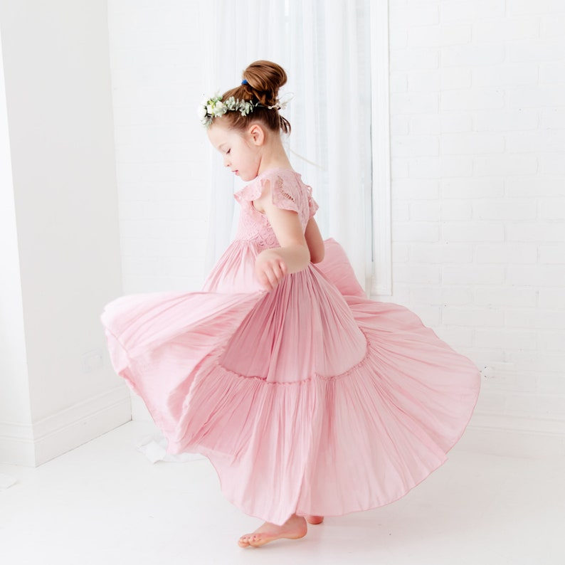 May include: A young girl wearing a pink, tiered dress with a floral crown spins around in a white room.
