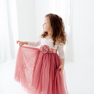 May include: A young girl wearing a white lace top with a pink tulle skirt and a pink flower embellishment. The skirt is flowing and the girl is looking to the side.