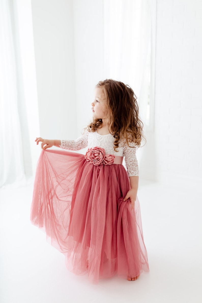 May include: A young girl wearing a white lace top with a pink tulle skirt and a pink flower embellishment. The skirt is flowing and the girl is looking to the side.