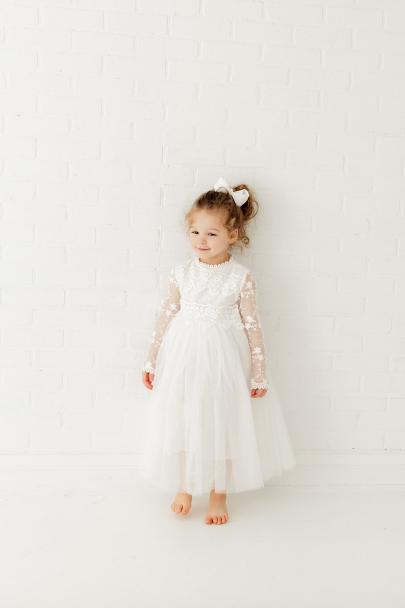 May include: A young girl wearing a white tulle dress with lace detailing and a white bow in her hair. She is standing in front of a white brick wall.