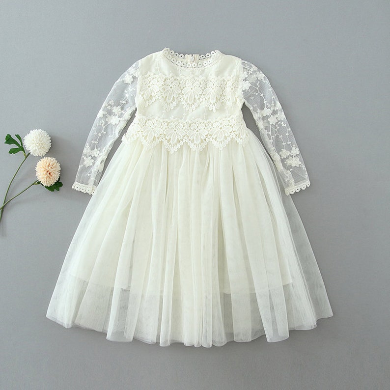 May include: A long-sleeved cream-colored dress featuring a round neckline, intricate floral lace detailing across the bodice and sleeves, and a layered tulle skirt. The garment is displayed alongside two small, pale decorative flowers.