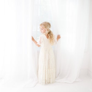 以下が含まれることがあります： 白いレースのドレスを着て、金の王冠のヘッドバンドをしている若い女の子が、白いカーテンの前に立って、窓の外を見ている。