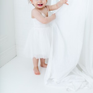 Puede incluir: Una ni&ntilde;a peque&ntilde;a con un vestido blanco de tul sonr&iacute;e mientras sostiene una cortina blanca en una habitaci&oacute;n blanca.