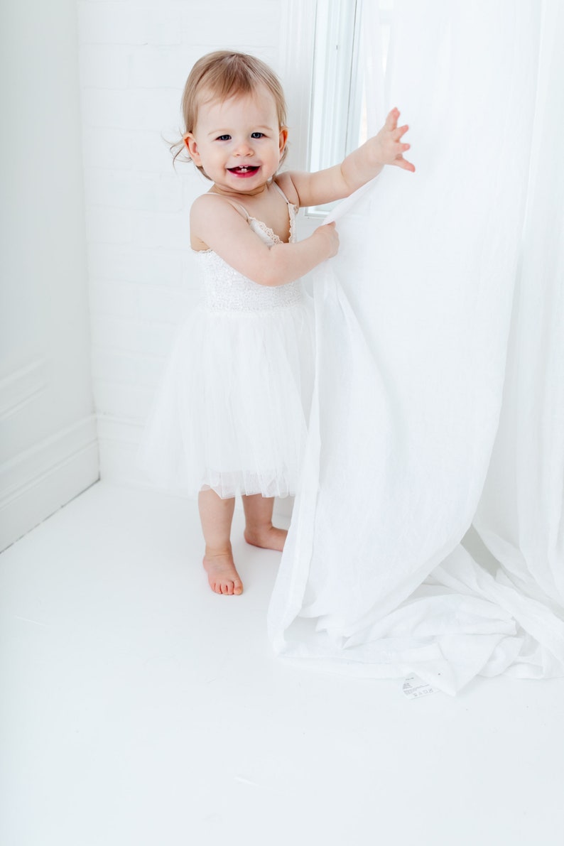 Puede incluir: Una ni&ntilde;a peque&ntilde;a con un vestido blanco de tul sonr&iacute;e mientras sostiene una cortina blanca en una habitaci&oacute;n blanca.