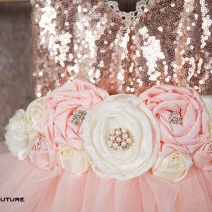 May include: A pink tulle tutu dress with a rose gold sequin bodice and a flower sash with pink and white fabric flowers. The sash has pearl and rhinestone embellishments.