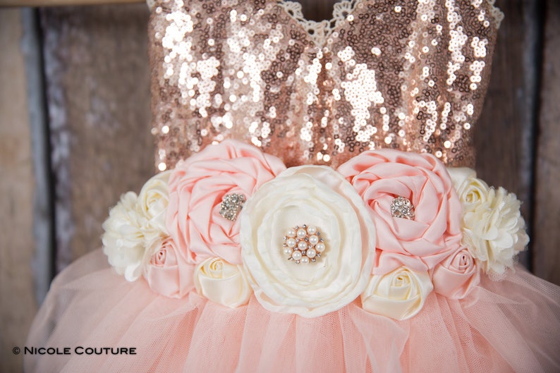 May include: A pink tulle tutu dress with a rose gold sequin bodice and a flower sash with pink and white fabric flowers. The sash has pearl and rhinestone embellishments.