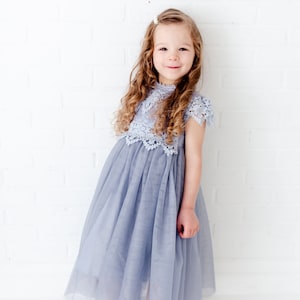 K&ouml;nnte beinhalten: Ein junges M&auml;dchen tr&auml;gt ein hellblaues T&uuml;llkleid mit einem Spitzenoberteil. Das Kleid hat einen hohen Ausschnitt und kurze &Auml;rmel. Das M&auml;dchen steht vor einer wei&szlig;en Backsteinwand.