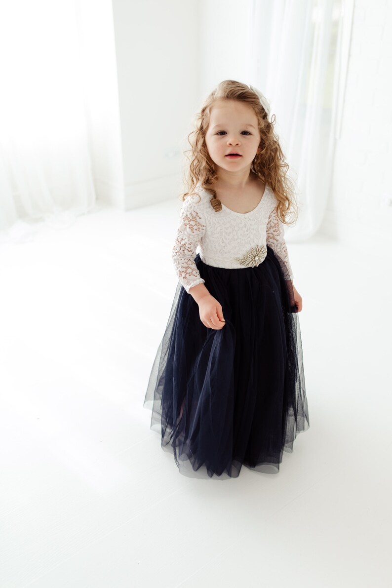 May include: A young girl wearing a white lace long-sleeved dress with a navy blue tulle skirt and a jeweled belt.