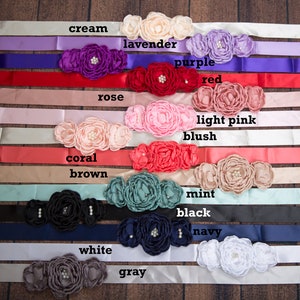 May include: A collection of satin sashes, each featuring a cluster of three fabric flowers with central pearl-like accents. The sashes are displayed in various colors labeled as cream, lavender, purple, red, rose, light pink, blush, coral, brown, mint, black, navy, white, and gray.