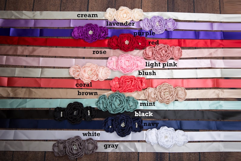 May include: A collection of satin sashes, each featuring a cluster of three fabric flowers with central pearl-like accents. The sashes are displayed in various colors labeled as cream, lavender, purple, red, rose, light pink, blush, coral, brown, mint, black, navy, white, and gray.