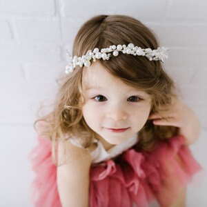 May include: A young girl with long brown hair wearing a pink tulle dress and a pearl and crystal headband.