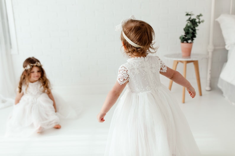 May include: Two young girls in white dresses. One girl is in the foreground, wearing a dress with lace sleeves and a tulle skirt. The other girl is seated, wearing a similar dress and a floral crown. The background is a white room with a small table and a plant.