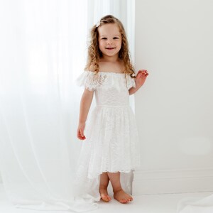 May include: A young girl wearing a white lace dress with an off-the-shoulder neckline and a high-low hemline. She is standing in front of a white curtain and smiling at the camera.