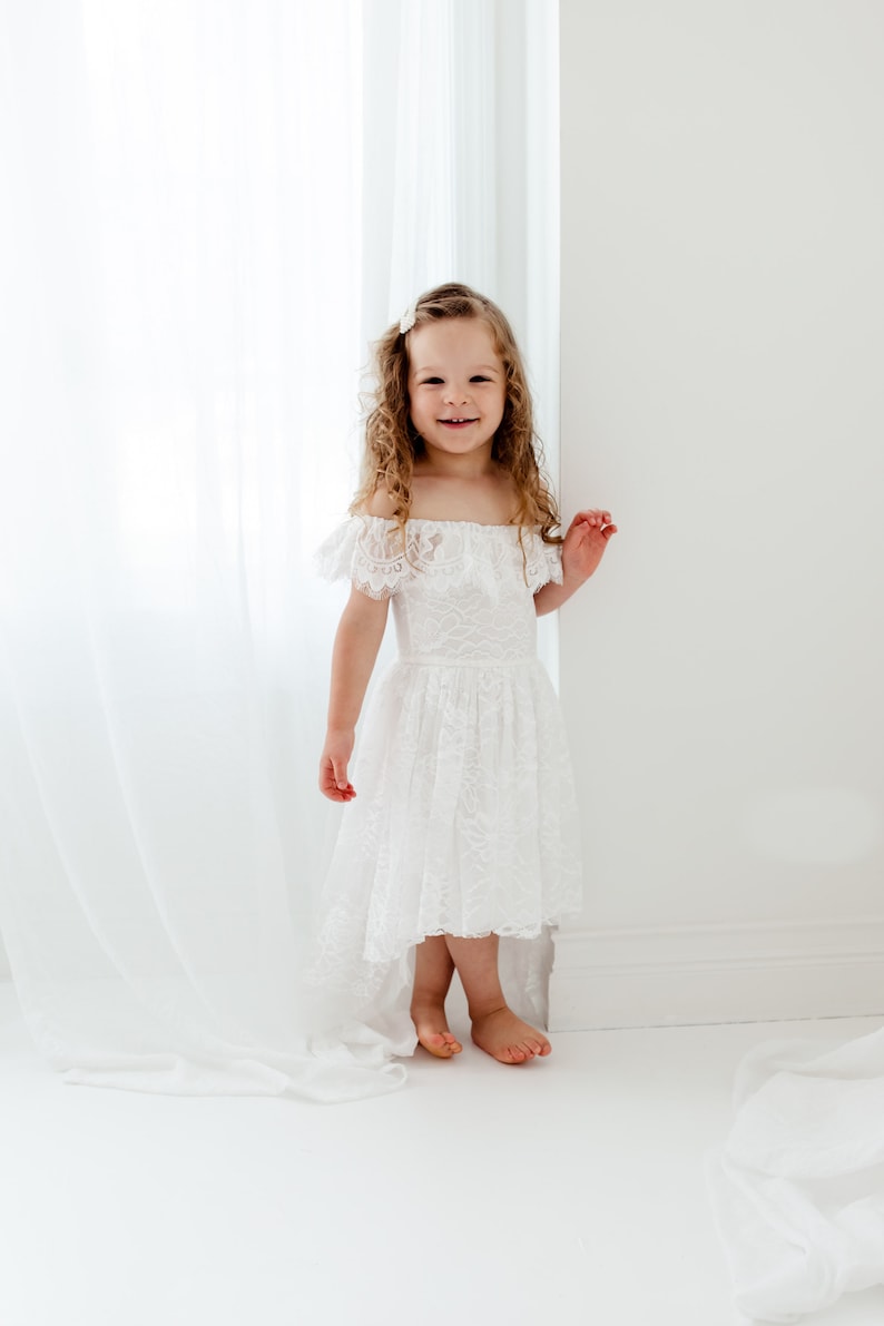 May include: A young girl wearing a white lace dress with an off-the-shoulder neckline and a high-low hemline. She is standing in front of a white curtain and smiling at the camera.