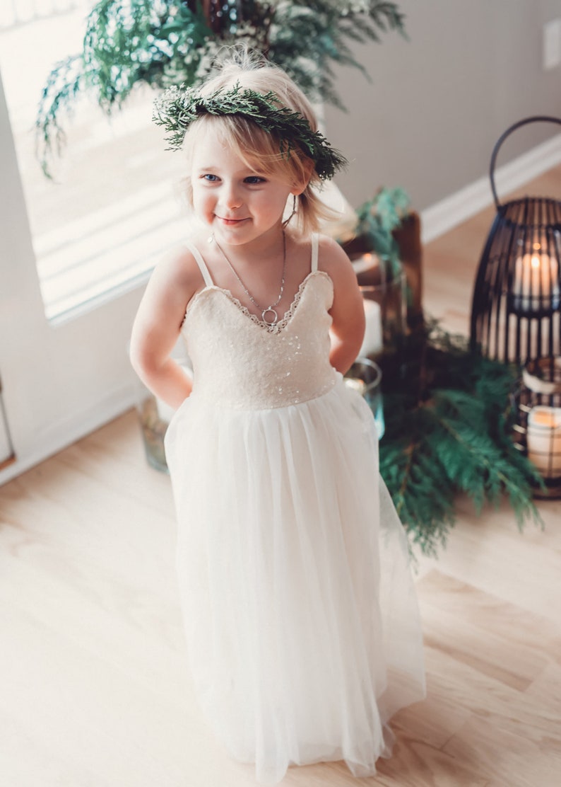 May include: A young girl wearing a white dress with a sequined bodice and a tulle skirt. She is wearing a green wreath in her hair and a silver necklace. The girl is standing in front of a window with greenery behind her.