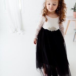 May include: A floor-length dress featuring a white long-sleeved lace bodice and a voluminous black tulle skirt. The waist is accented with a decorative silver-toned brooch featuring clear crystals. The dress has a scoop neckline and long sleeves.