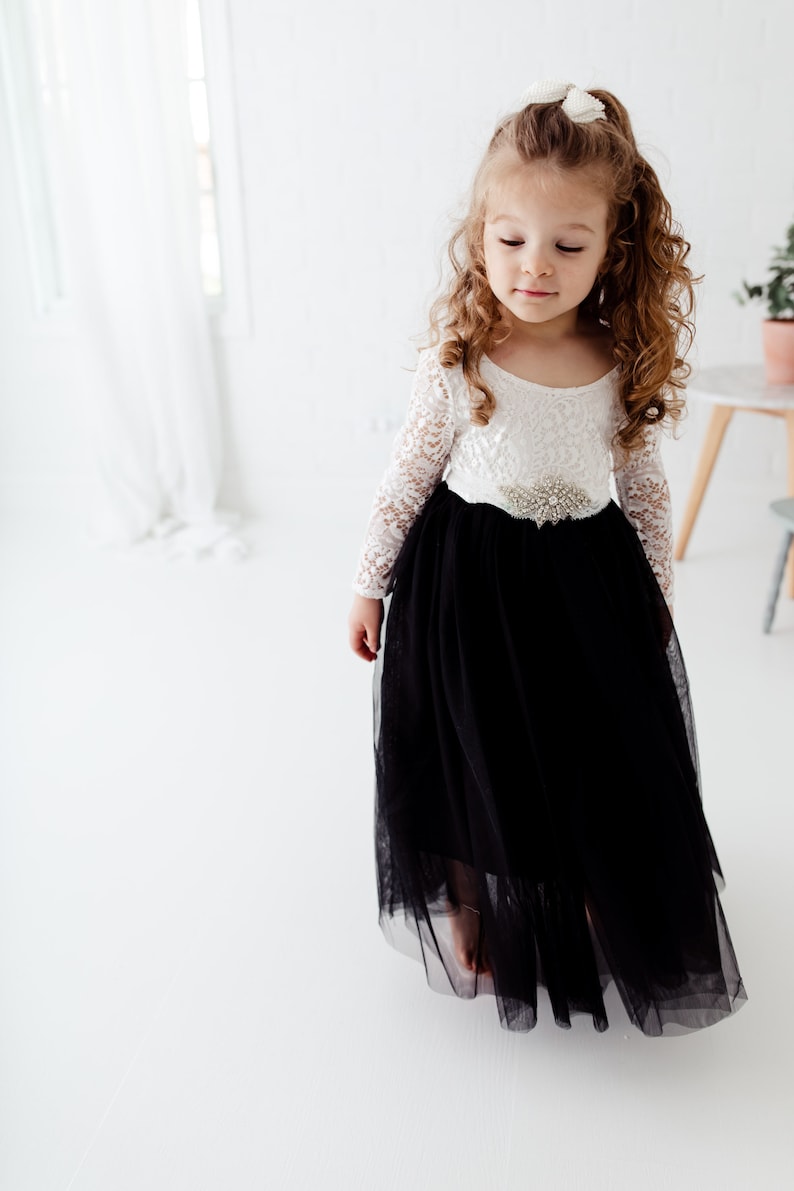 May include: A floor-length dress featuring a white long-sleeved lace bodice and a voluminous black tulle skirt. The waist is accented with a decorative silver-toned brooch featuring clear crystals. The dress has a scoop neckline and long sleeves.