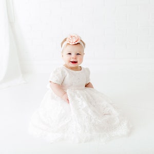 May include: A white lace dress with short sleeves and a full skirt, worn with a matching white headband featuring a large, pale peach-colored fabric flower. The garment has a textured floral pattern throughout and a delicate scalloped hemline.