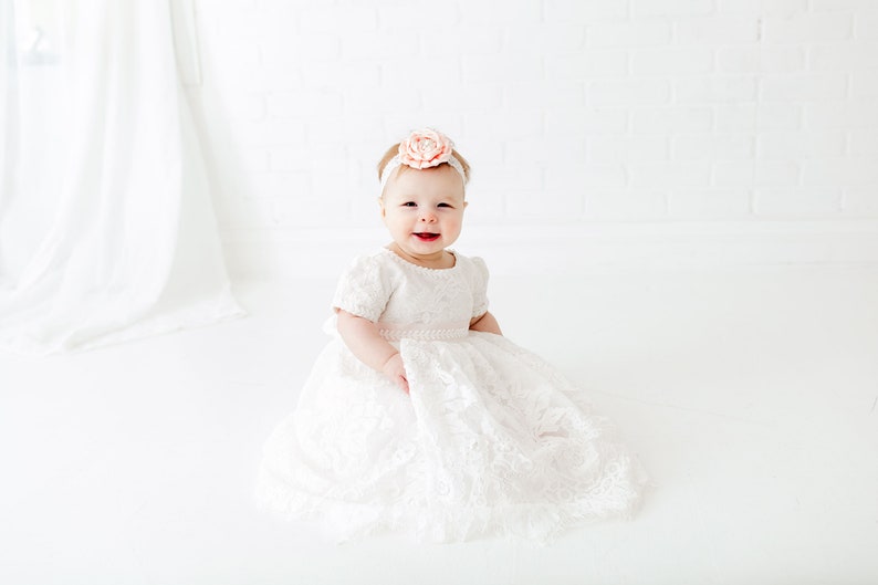 May include: A white lace dress with short sleeves and a full skirt, worn with a matching white headband featuring a large, pale peach-colored fabric flower. The garment has a textured floral pattern throughout and a delicate scalloped hemline.